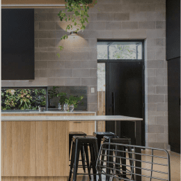 The modern kitchen features a wooden island, black stools, and a metal chair. A small plant graces the counter while a larger one hangs on the wall. With Austral Masonry Blocks framing the sleek black appliances the space exudes contemporary charm. Doorway in the background.